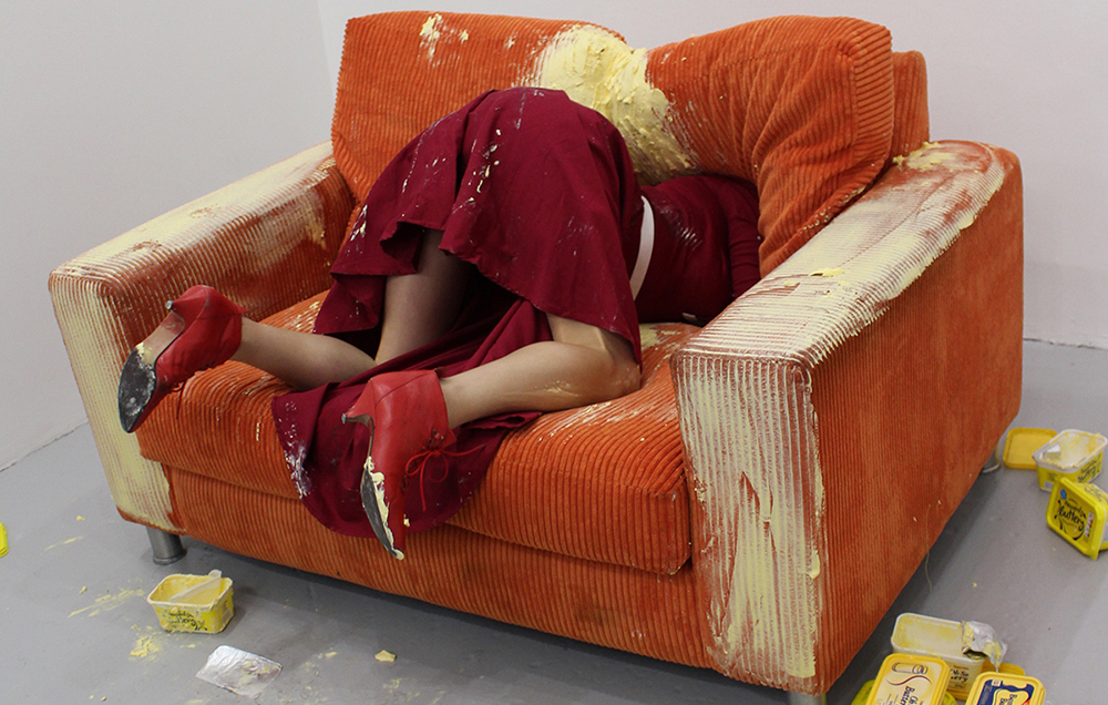 a figure dressed in a red dress and shows dives head first into an orange sofa that is covered in butter, with tubs of butter lying on the floor beside them