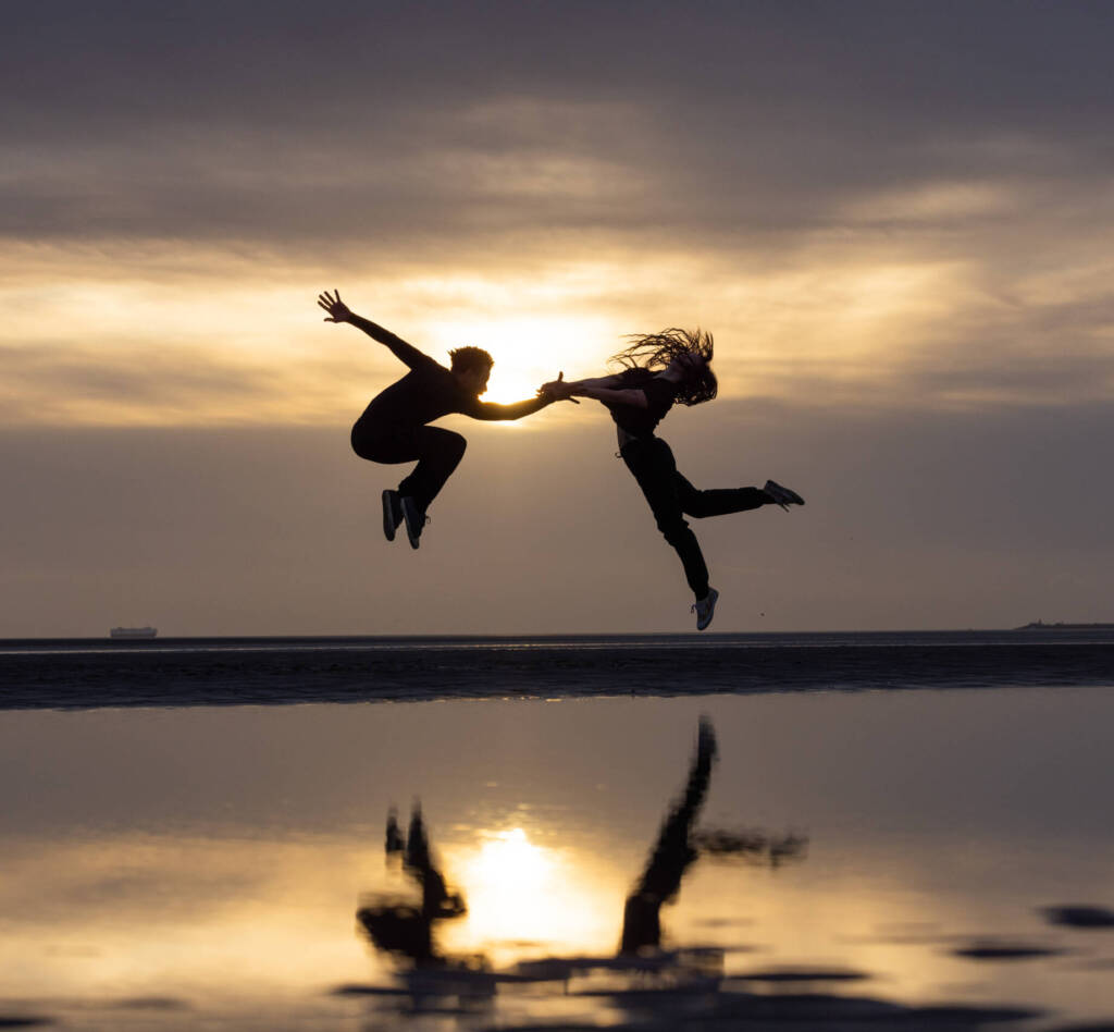 Dancers Vitor Barboza da Silva and Inez Berdychowska are seen in silhouette, jumping on a beach, adopting dramatic poses. Behind them a cloudy dawn sky is visible. Their silhouettes are reflected in a pool of water beneath them.