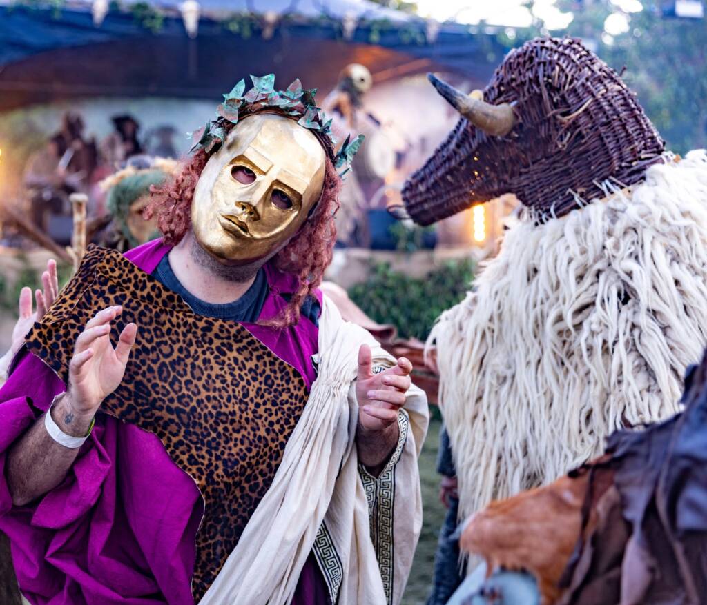 Two people in vibrant costumes pose for the camera. One wears a golden mask with ivy leaves an da curly red wig, the other has a woven mask with horns and a woolly collar.