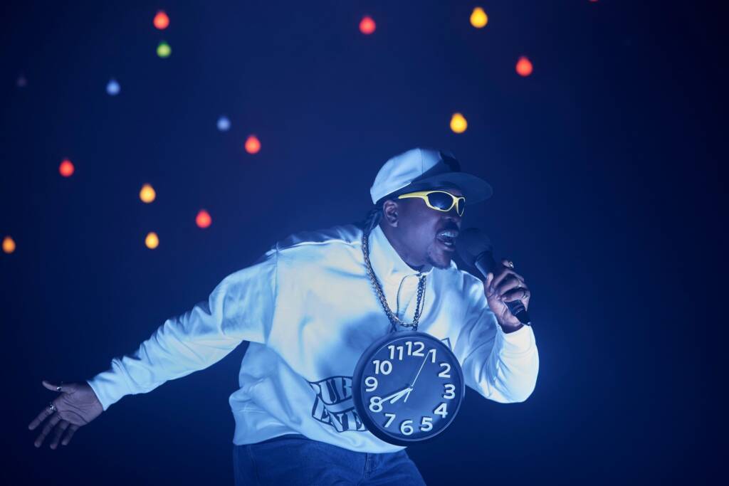 Artist Ahmed Karim Tamu captured in performance. He sings into a microphone with his other arm extended behind his dramatically. He wears a white jack and cap with a clock on a chain and a pair of neon yellow sunglasses