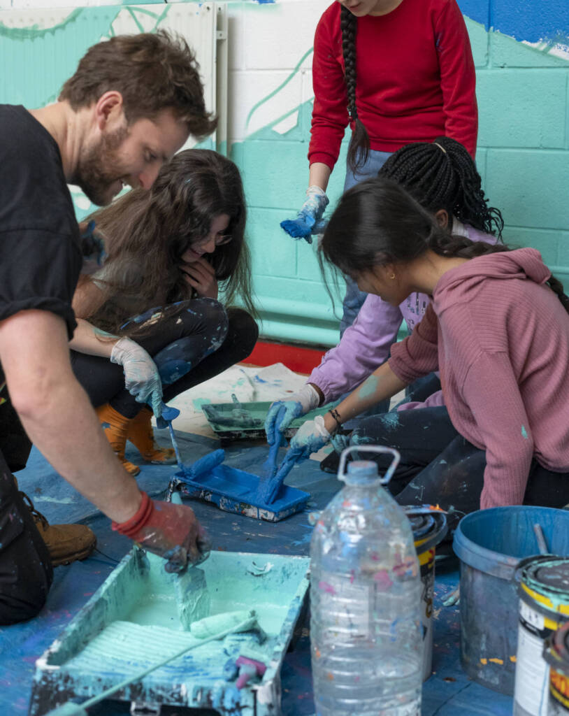 A group of young students reach into paint trays with their brushes as they paint a large mural on the wall behind them
