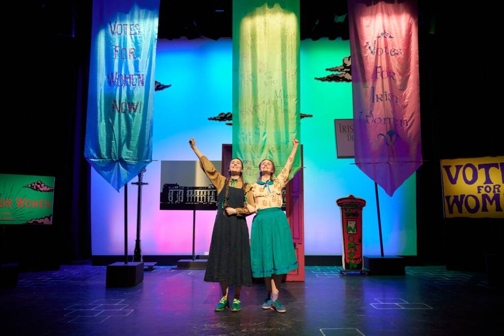two actresses stand on stage with their fist raised to the sky in triumph. Behind them are various posters for womens rights and votes for women.