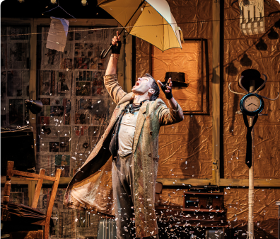A performer stands on stage holding an umbrella and dramatically raises their arms to the sky. Confetti swirls around them.